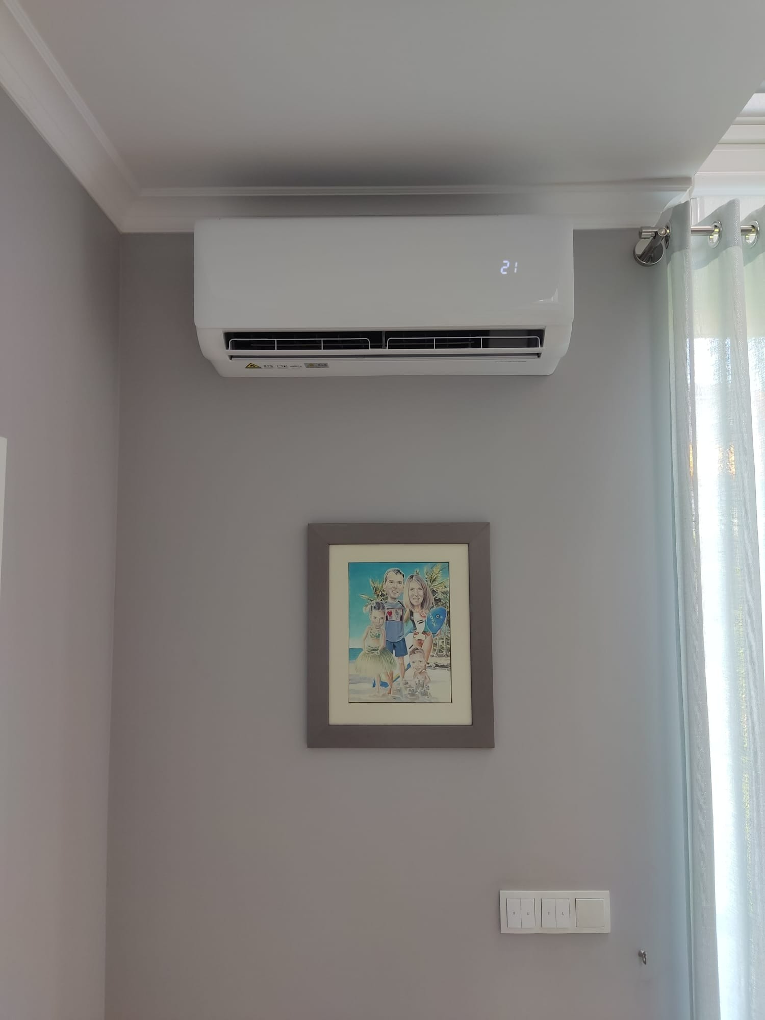 Biały klimatyzator ścienny zamontowany w pokoju z szarą ścianą, pod nim wisi obrazek w ramce, a obok okno z zasłoną.