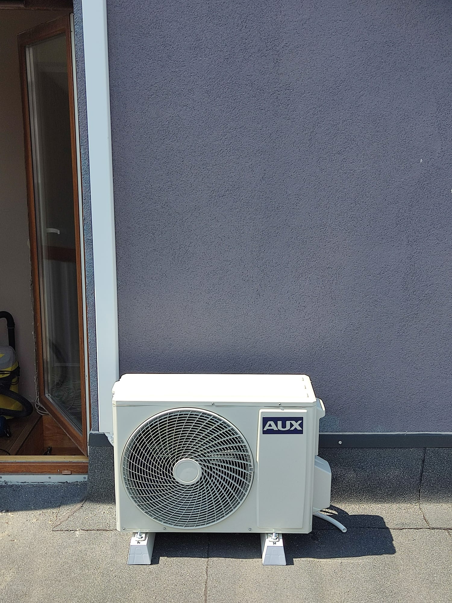 Zewnętrzna jednostka klimatyzacji AUX zamontowana na dachu budynku z szarą, teksturowaną ścianą i otwartym oknem w tle.