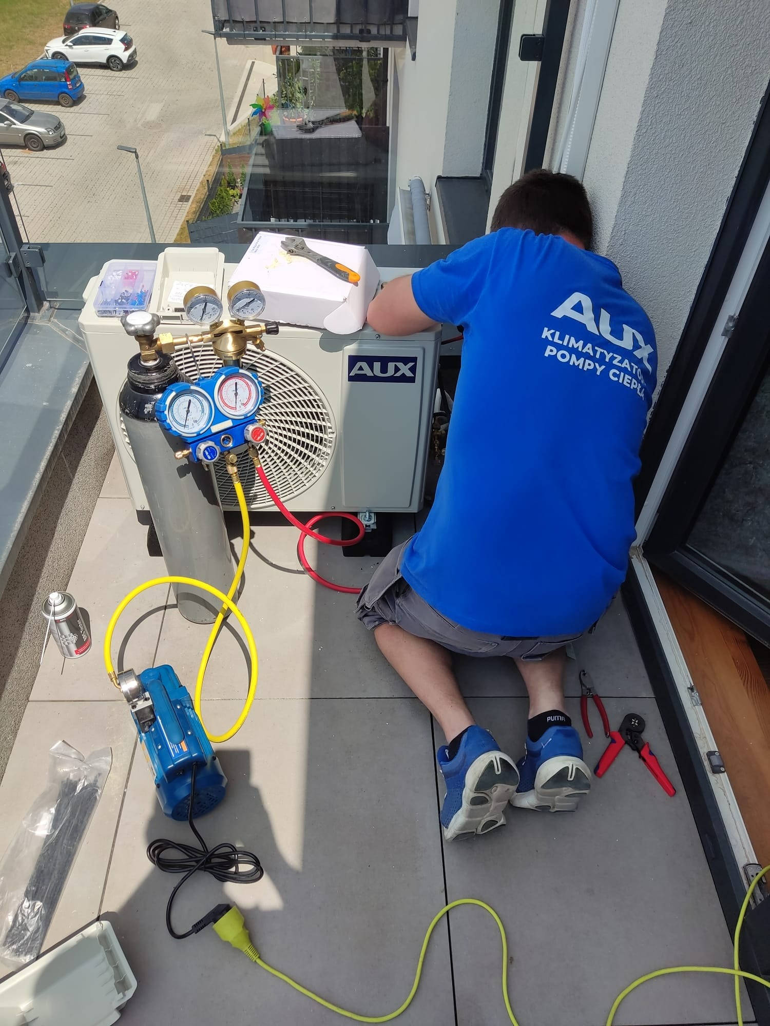 Instalator w niebieskiej koszulce z logo AUX podłącza manometry do klimatyzatora na balkonie, widoczne narzędzia i butla z gazem.