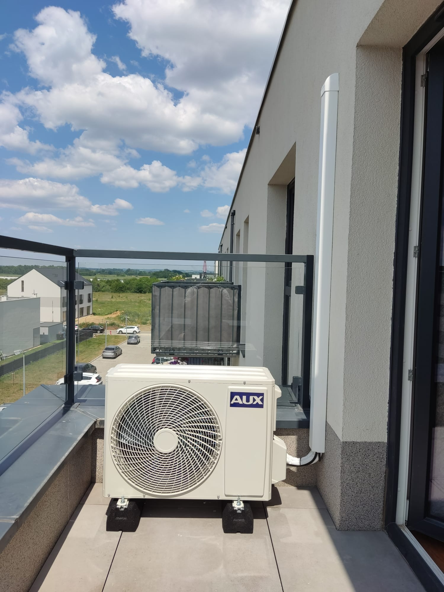Zewnętrzna jednostka klimatyzacji marki AUX zamontowana na balkonie w nowoczesnym budynku mieszkalnym, widok na okolicę z błękitnym niebem i chmurami.