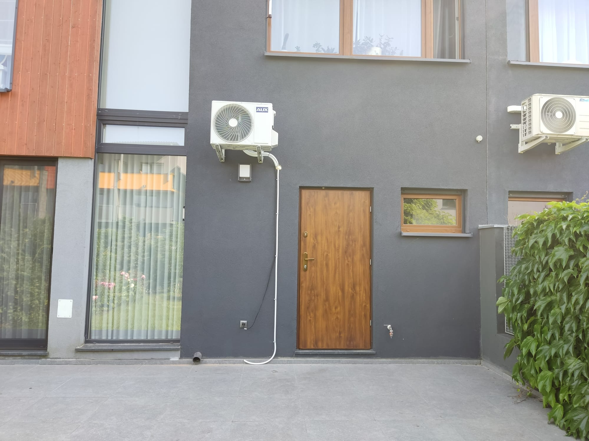 Szara fasada domu z dwoma zamontowanymi klimatyzatorami typu split, widoczne okna i drewniane drzwi wejściowe.