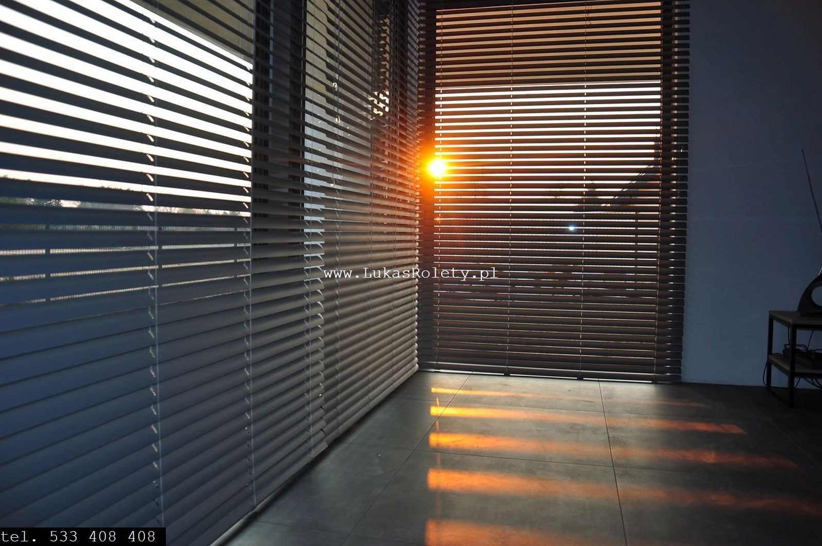 Zasłonięte żaluzje poziome w nowoczesnym wnętrzu, z promieniami zachodzącego słońca wpadającymi przez szpary, tworzącymi refleksy na podłodze.