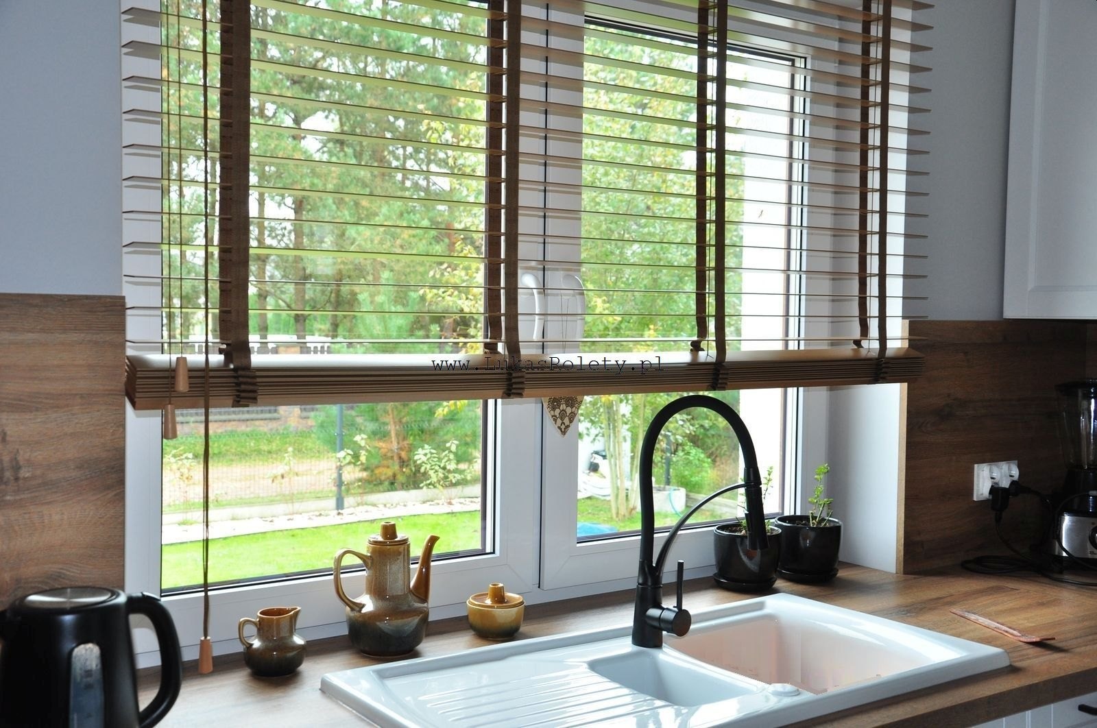 Kuchnia z drewnianymi żaluzjami w oknie, widok na ogród, czajnik i filiżanki na blacie, czarna bateria kuchenna.