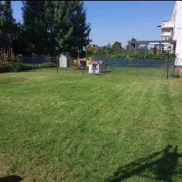 Rozległy trawnik przed nowoczesnym domem z placem zabaw w tle, świeżo skoszona trawa z widocznymi śladami po kosiarce.