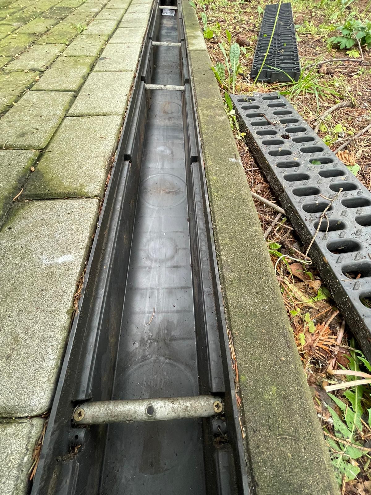 Otwarty, czarny kanał odwadniający z metalowymi poprzeczkami, ułożony wzdłuż chodnika z szarych płyt betonowych. Obok leżą zdjęte, czarne kratki odwadniające, a w tle widać roślinność.