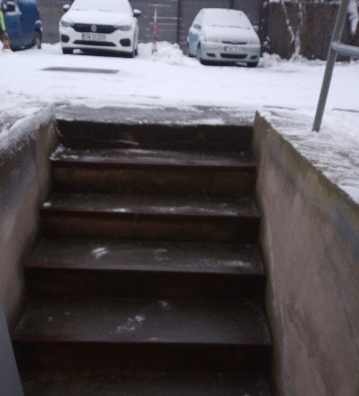Zewnętrzne schody prowadzące w dół, pokryte śniegiem, z samochodami zaparkowanymi w tle.