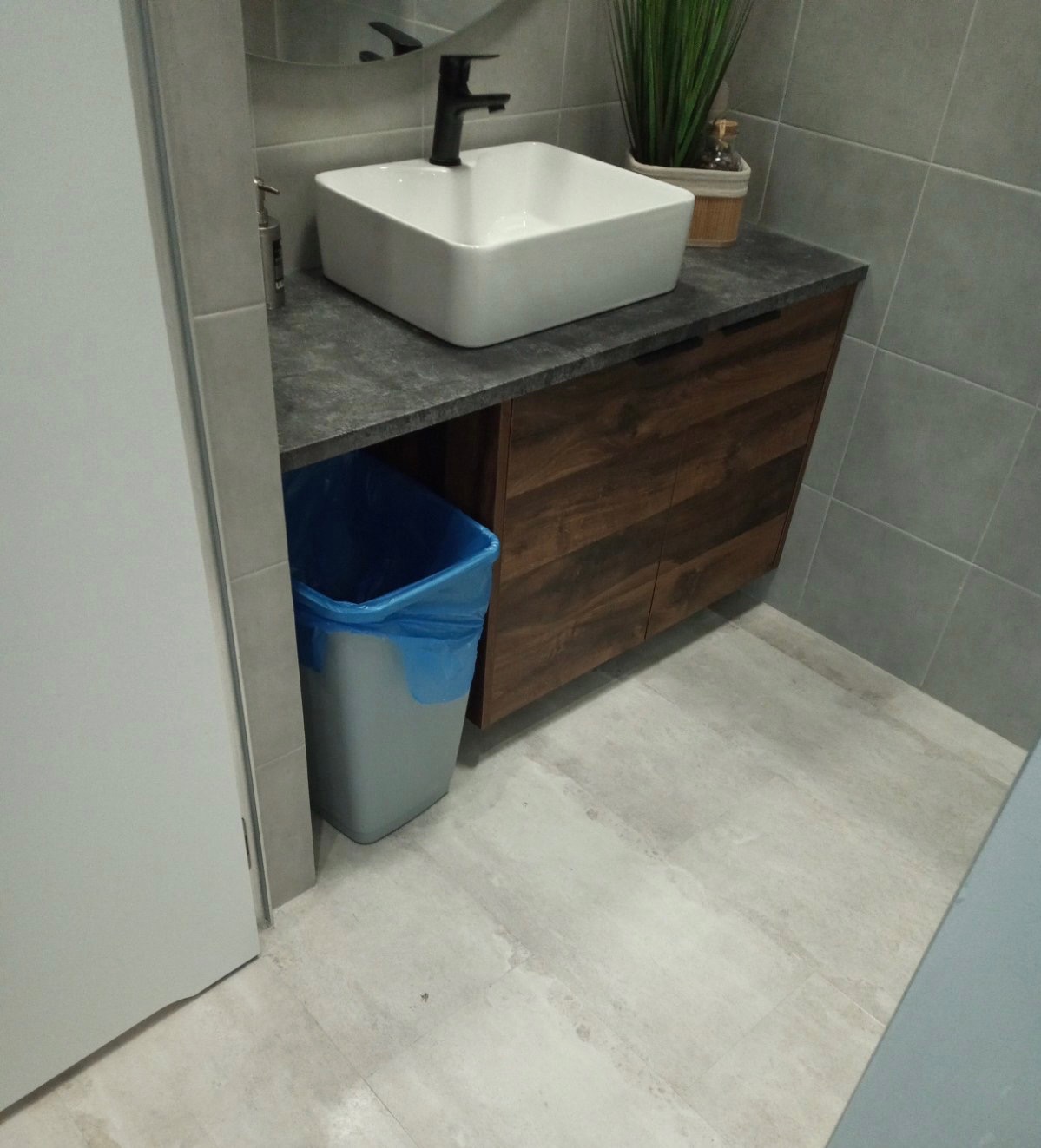 Nowoczesna łazienka z kwadratową umywalką, czarną armaturą i szafką z drewnopodobnym frontem. Szary kosz na śmieci z niebieską wkładką stoi obok szafki, na podłodze płytki imitujące beton.