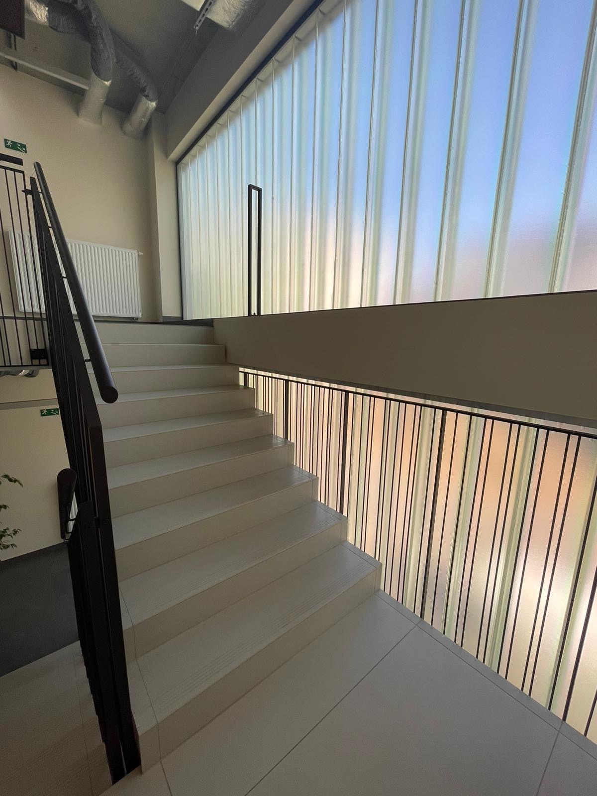 Białe schody w nowoczesnym budynku, z czarną metalową balustradą i ścianą z pionowych paneli przepuszczających światło.