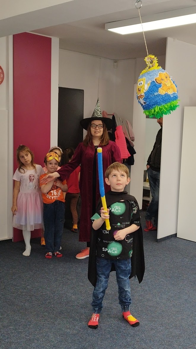 Chłopiec w stroju superbohatera trzymający pałkę do rozbijania piniaty w kształcie kuli ziemskiej z postaciami z bajek, w tle dzieci i kobieta w stroju czarownicy.