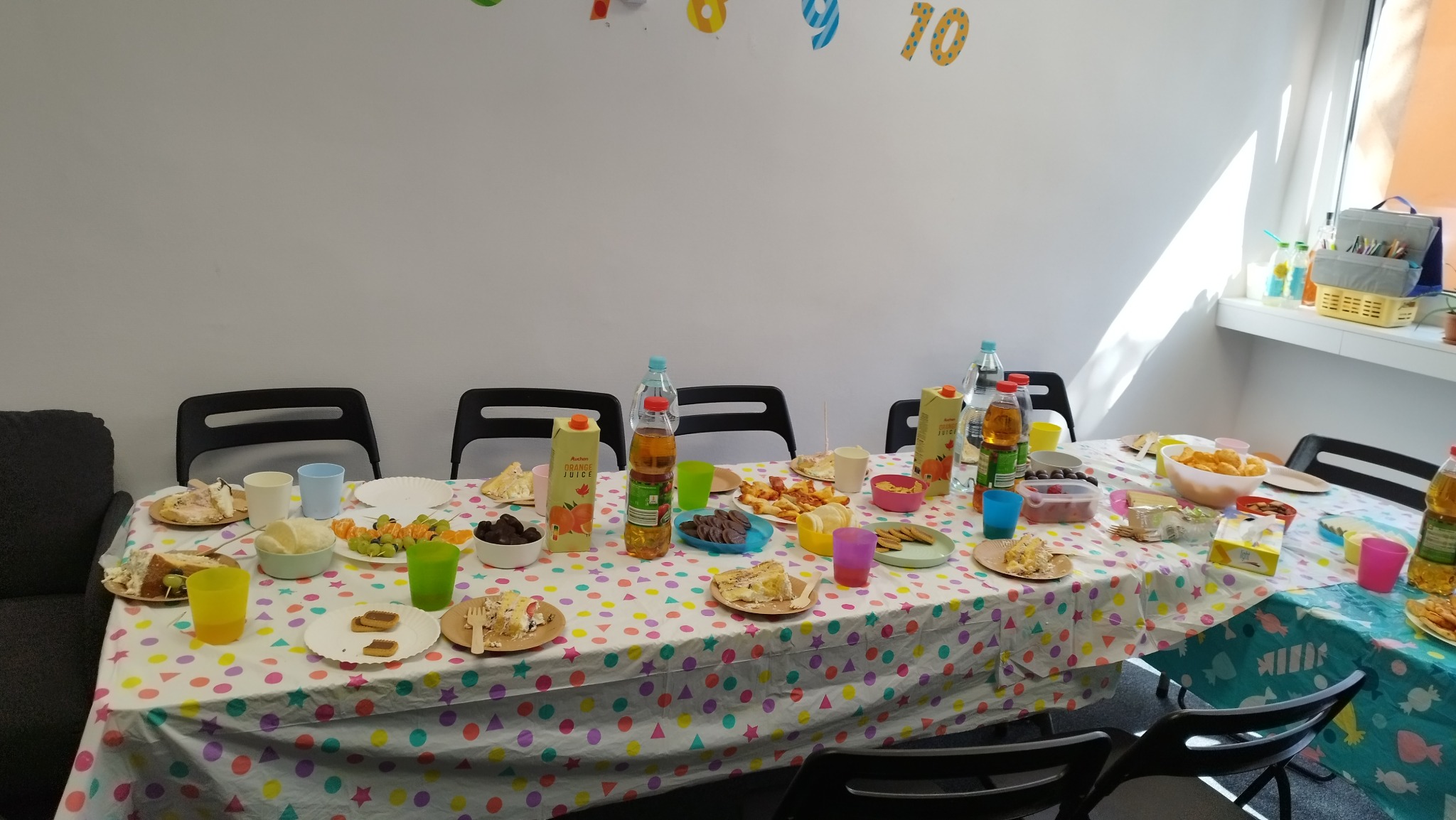 Stół nakryty kolorowym obrusem w kropki, zastawiony talerzami z jedzeniem, napojami i przekąskami, przygotowany na przyjęcie urodzinowe dla dzieci, z dekoracjami cyfrowymi na ścianie.