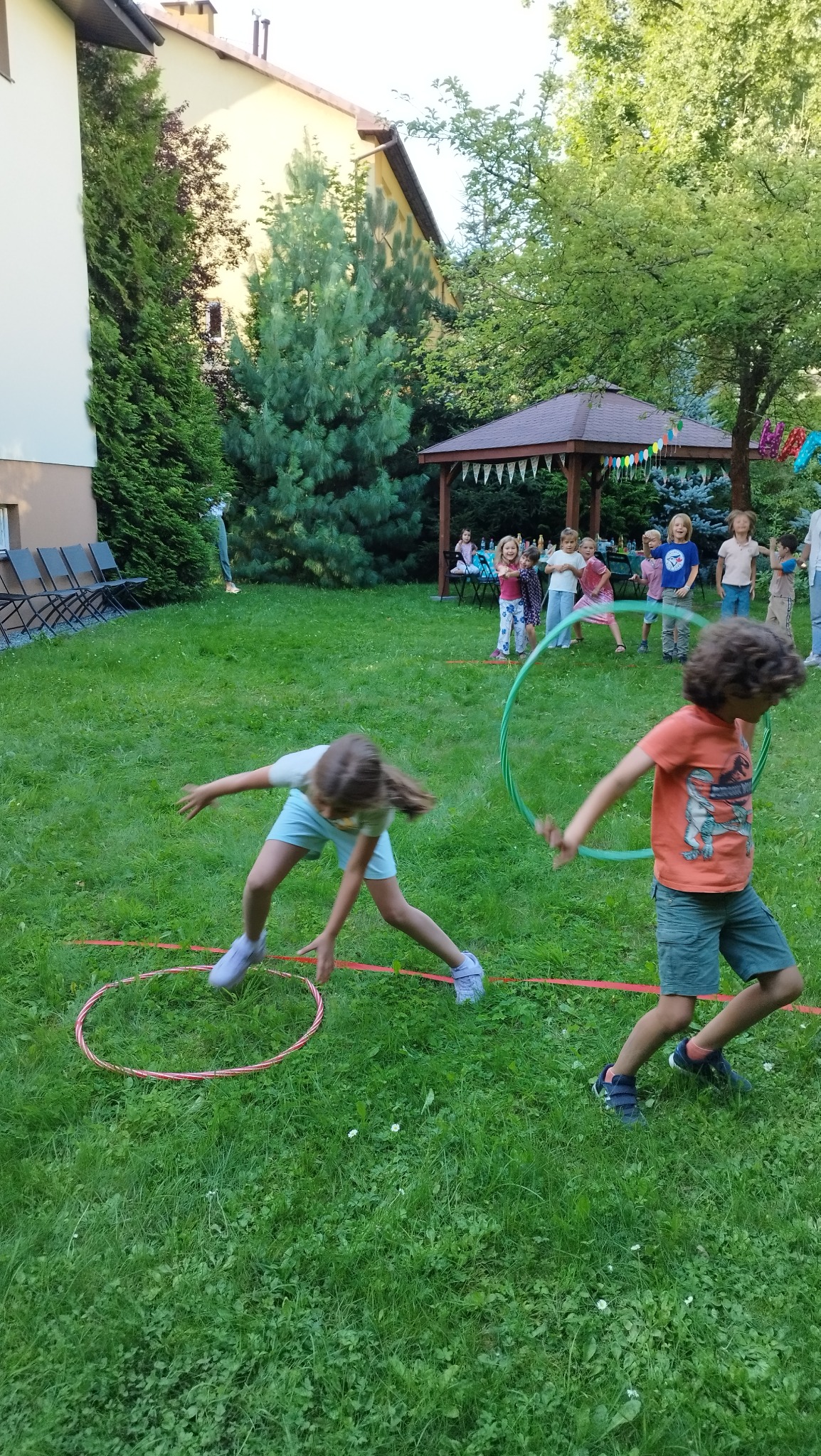 Dzieci bawią się hula-hop na trawie podczas imprezy plenerowej, w tle widoczny altana i grupa dzieci.