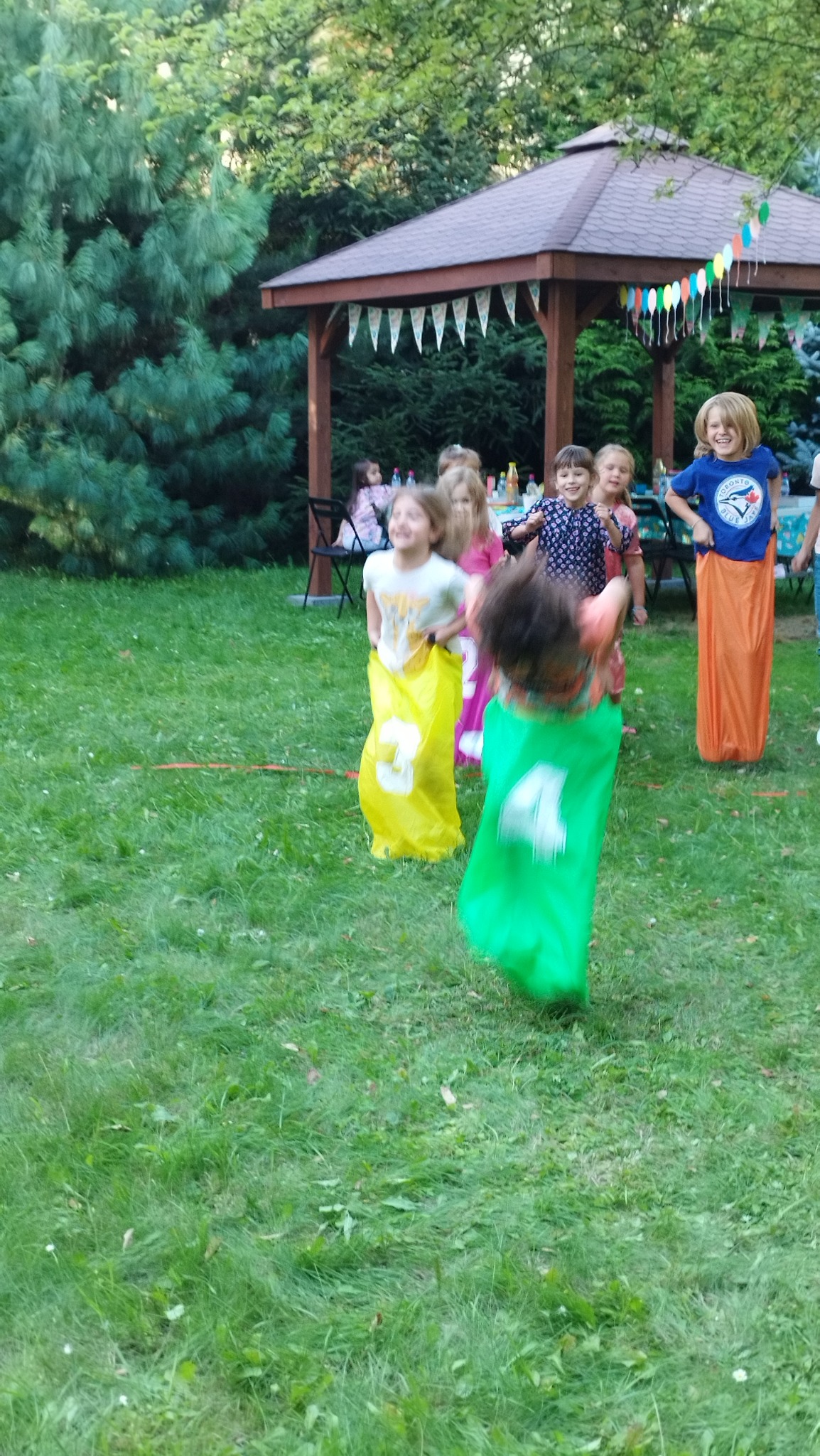 Radosne dzieci biorące udział w wyścigu w workach podczas imprezy plenerowej w Łodzi, z dekoracyjnym altanką w tle.