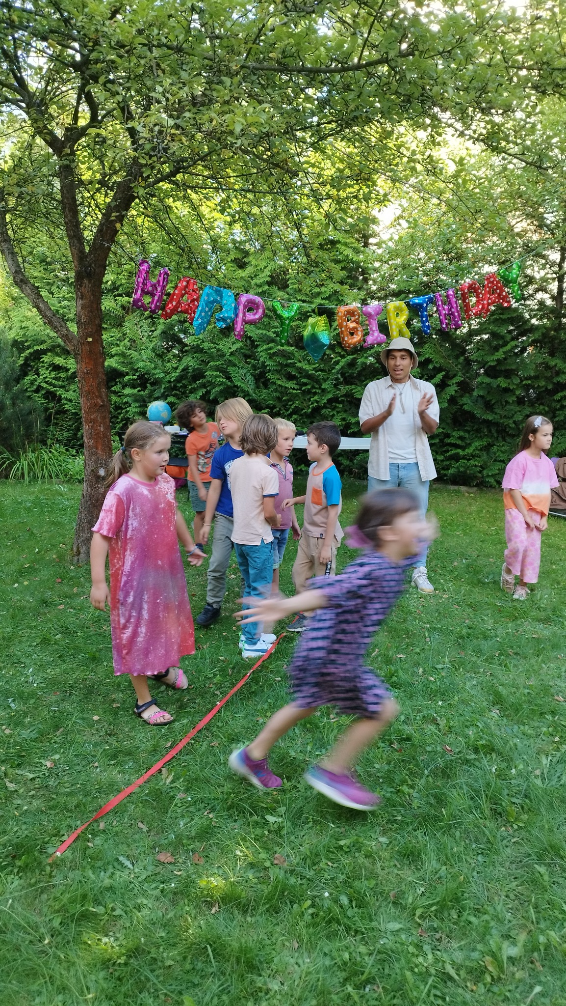 Dzieci na urodzinach w ogrodzie, biegają po trawie, nad nimi balonowy napis 'Happy Birthday', animator w kapeluszu klaszcze.