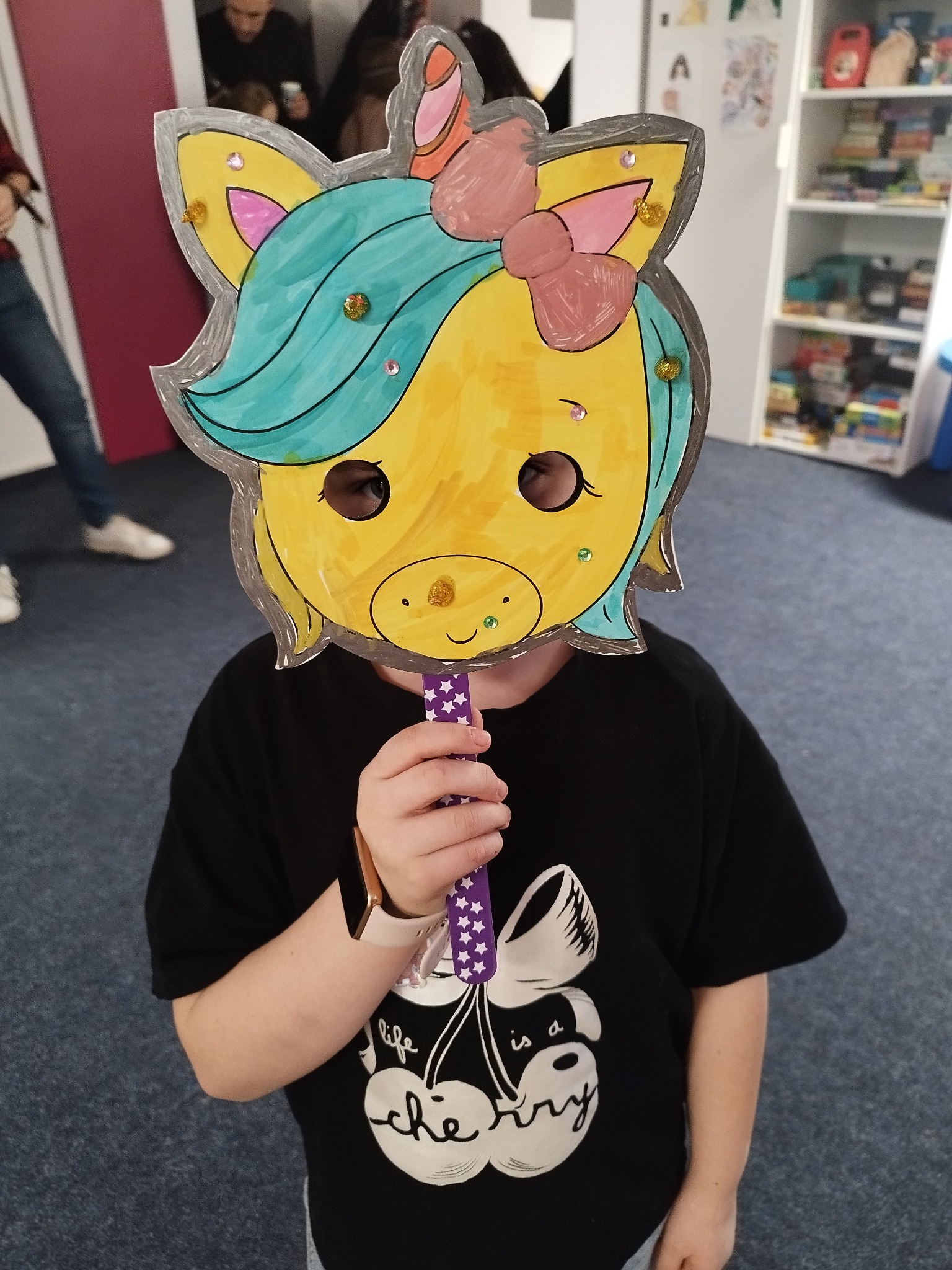 Dziecko trzyma samodzielnie wykonaną, kolorową maskę jednorożca na patyku, ozdobioną brokatem i cekinami, z widocznymi oczami dziecka przez wycięte otwory.