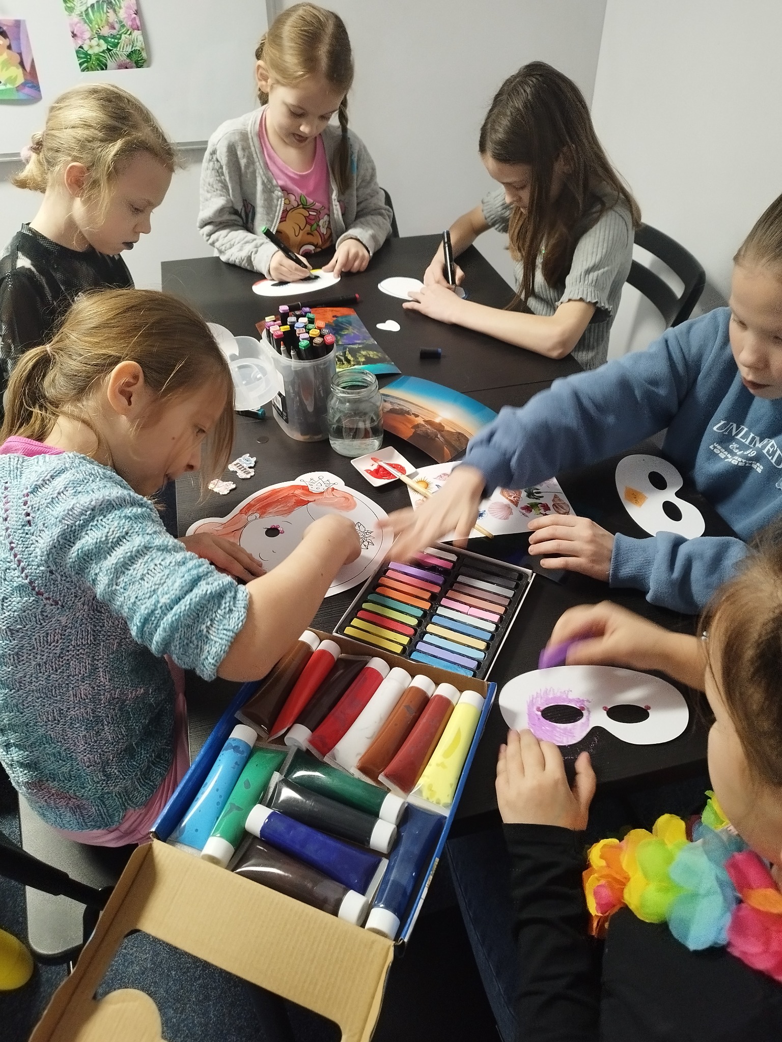 Grupa dzieci siedzi przy czarnym stole, dekorując papierowe maski karnawałowe farbami, kredkami pastelowymi i mazakami; widoczna tęczowa girlanda z kwiatów.
