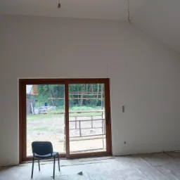 Wnętrze w trakcie wykańczania: świeżo pomalowane ściany na biało, surowe podłoże zabezpieczone folią, widoczne przewody elektryczne zwisające z sufitu, okno przesuwne z widokiem na zewnątrz...