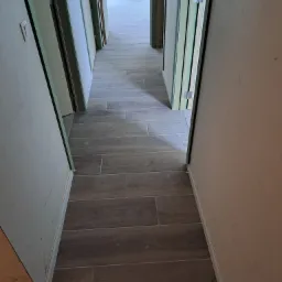 Długi korytarz w trakcie remontu z ułożonymi panelami podłogowymi imitującymi drewno, widoczne surowe ściany i ościeżnice drzwi.