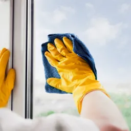 Mycie okna od wewnątrz: dłoń w żółtej rękawicy z niebieską ściereczką na tle błękitnego nieba widocznego przez szybę.