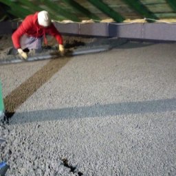 LbM wylewki maszynowe - Pracownik w czerwonej bluzie wyrównuje świeżo wylaną, szarą posadzkę w pomieszczeniu z drewnianym stropem, widoczny zielony słup i folia ochronna na podłodze.