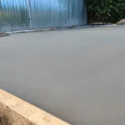 Wygładzona powierzchnia świeżo wylanego jastrychu cementowego, widoczna zacieraczka mechaniczna, szalunek z desek i blaszana ściana w tle.