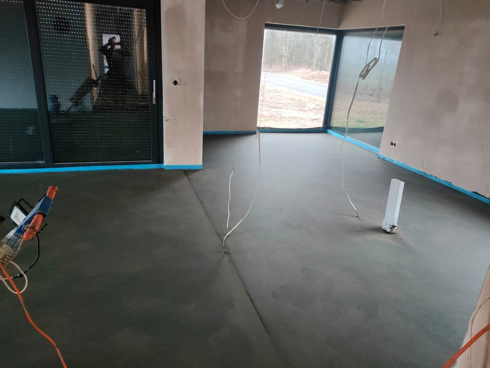 Wnętrze w stanie surowym z wylaną, szarą posadzką jastrychową, zabezpieczoną niebieską taśmą przy ścianach i otworach okiennych, widoczne kable wystające z podłogi oraz narzędzia w rogu pomieszczenia.