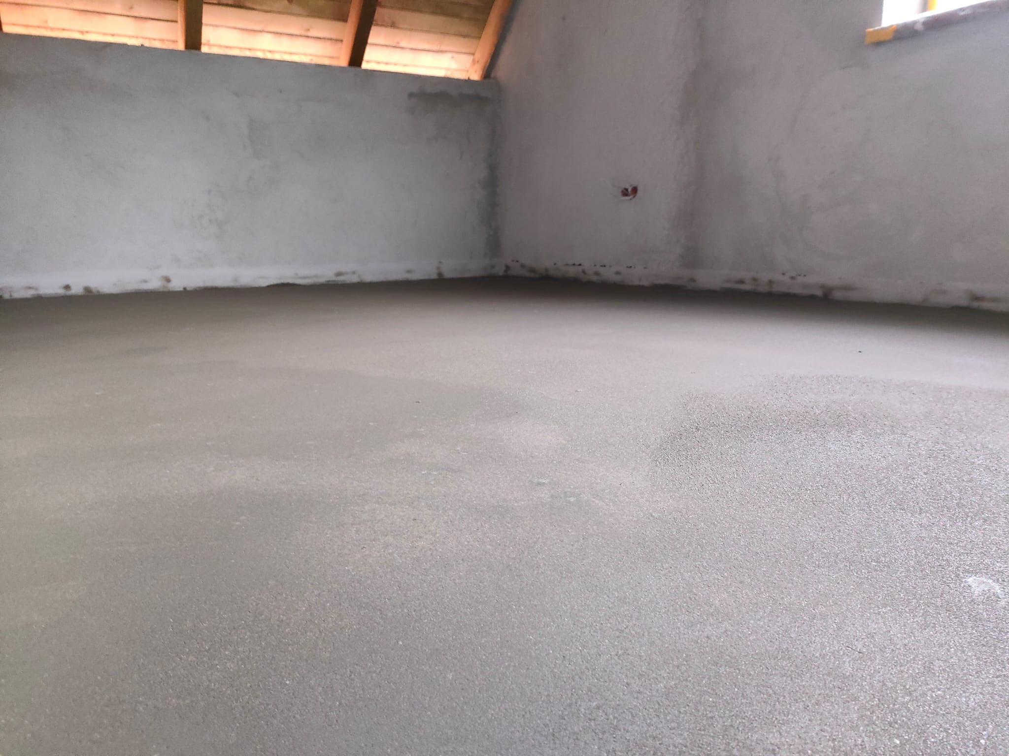 Świeżo wylana, gładka, szara posadzka jastrychowa w surowym wnętrzu, widoczne ślady wilgoci, drewniany strop i fragment okna.