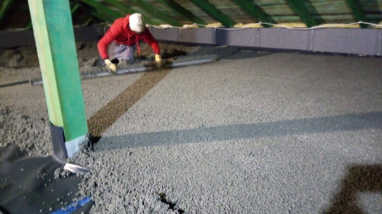 Pracownik w czerwonej bluzie wyrównuje świeżo wylaną, szarą posadzkę w pomieszczeniu z drewnianym stropem, widoczny zielony słup i folia ochronna na podłodze.