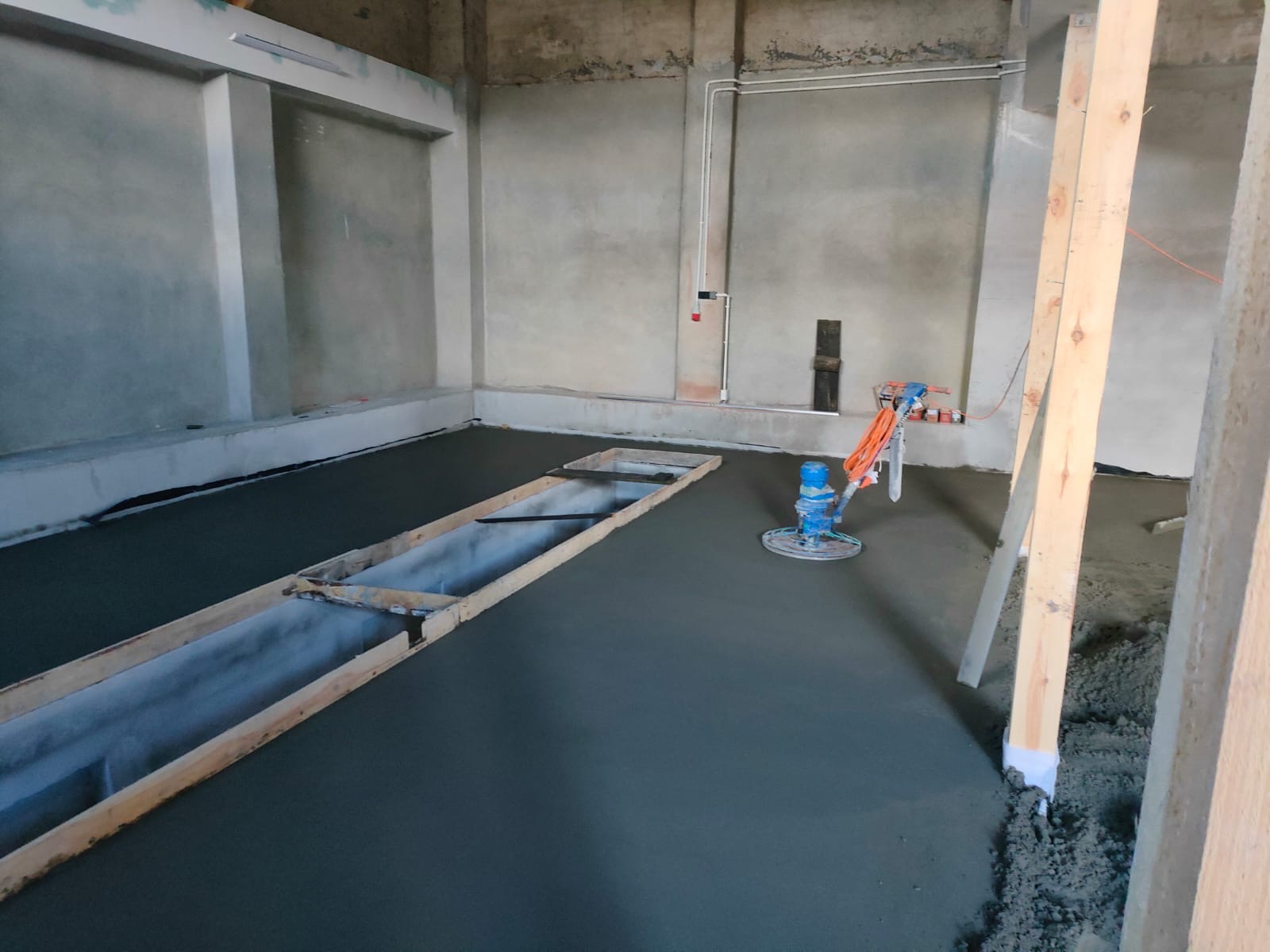 Świeżo wylana, szara posadzka jastrychowa w surowym wnętrzu, z widoczną maszyną do zacierania i szalunkiem z desek.