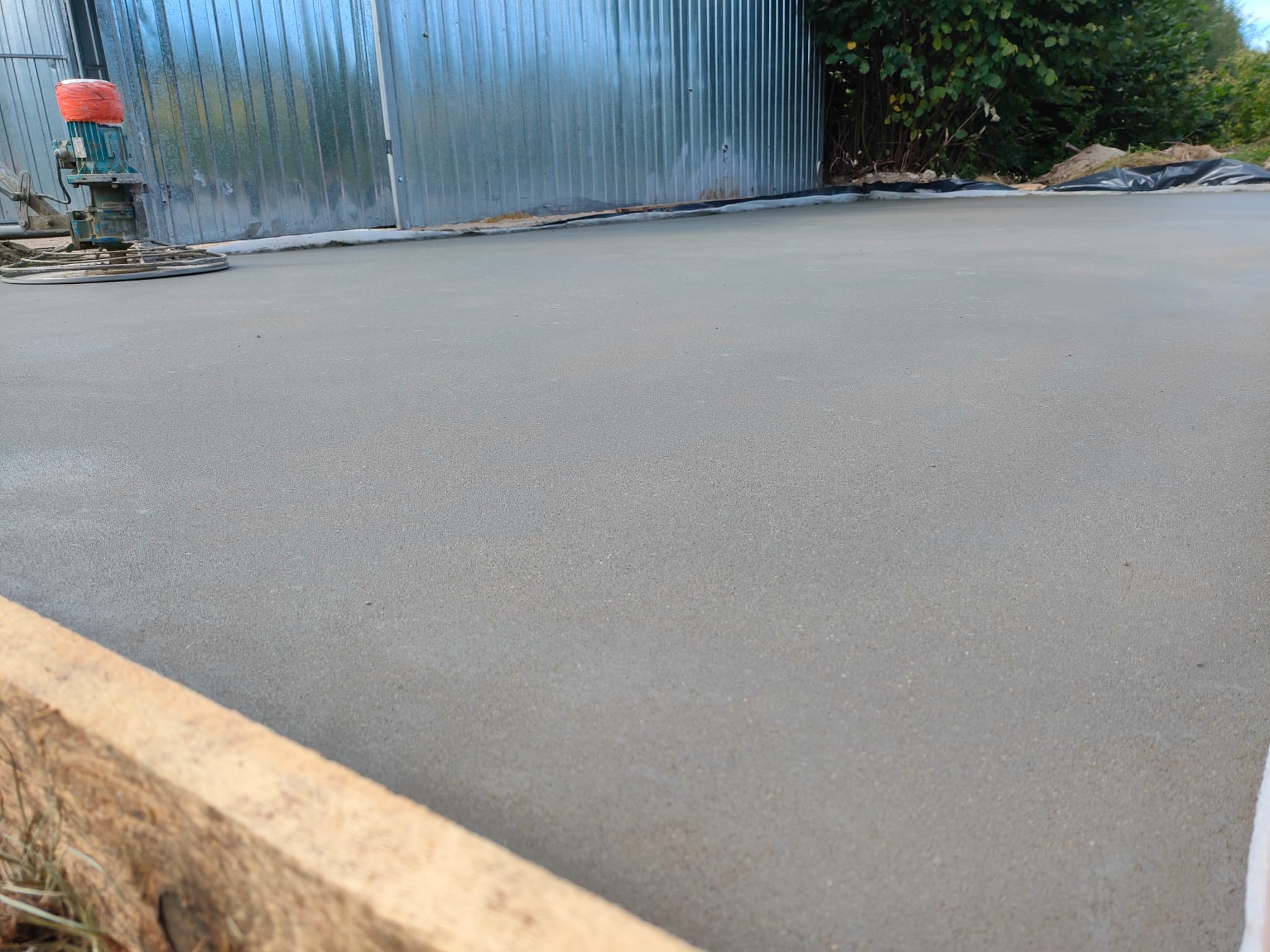 Wygładzona powierzchnia świeżo wylanego jastrychu cementowego, widoczna zacieraczka mechaniczna, szalunek z desek i blaszana ściana w tle.