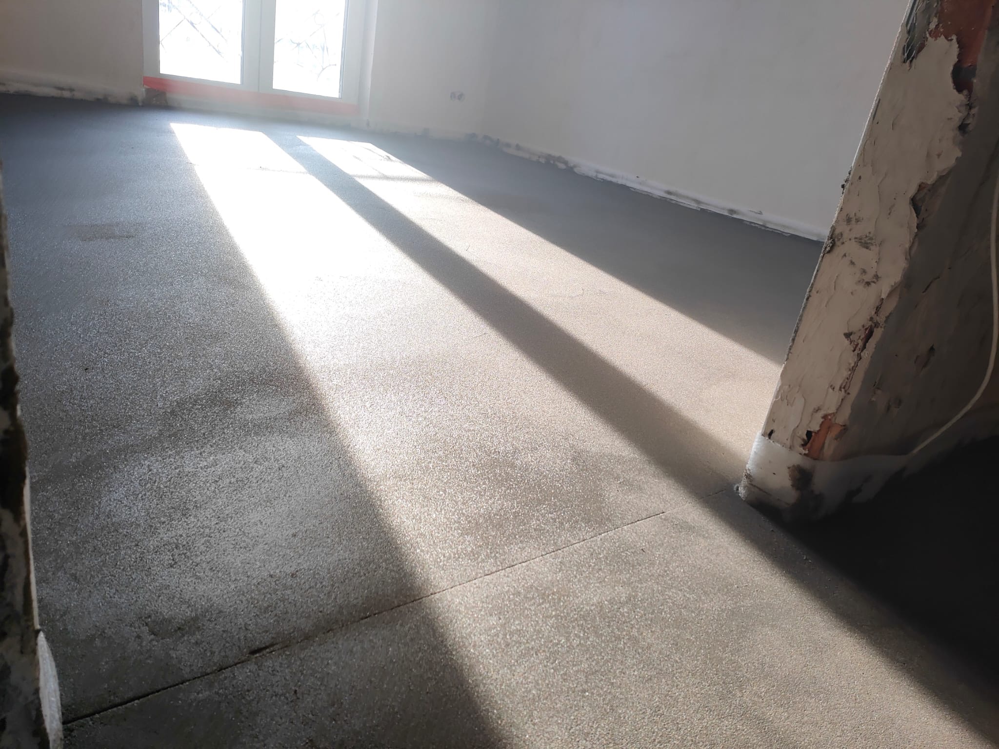 Świeżo wylana, szara posadzka w jasnym pomieszczeniu z dużym oknem, na której widoczne są promienie słoneczne i fragment brudnej drabiny.