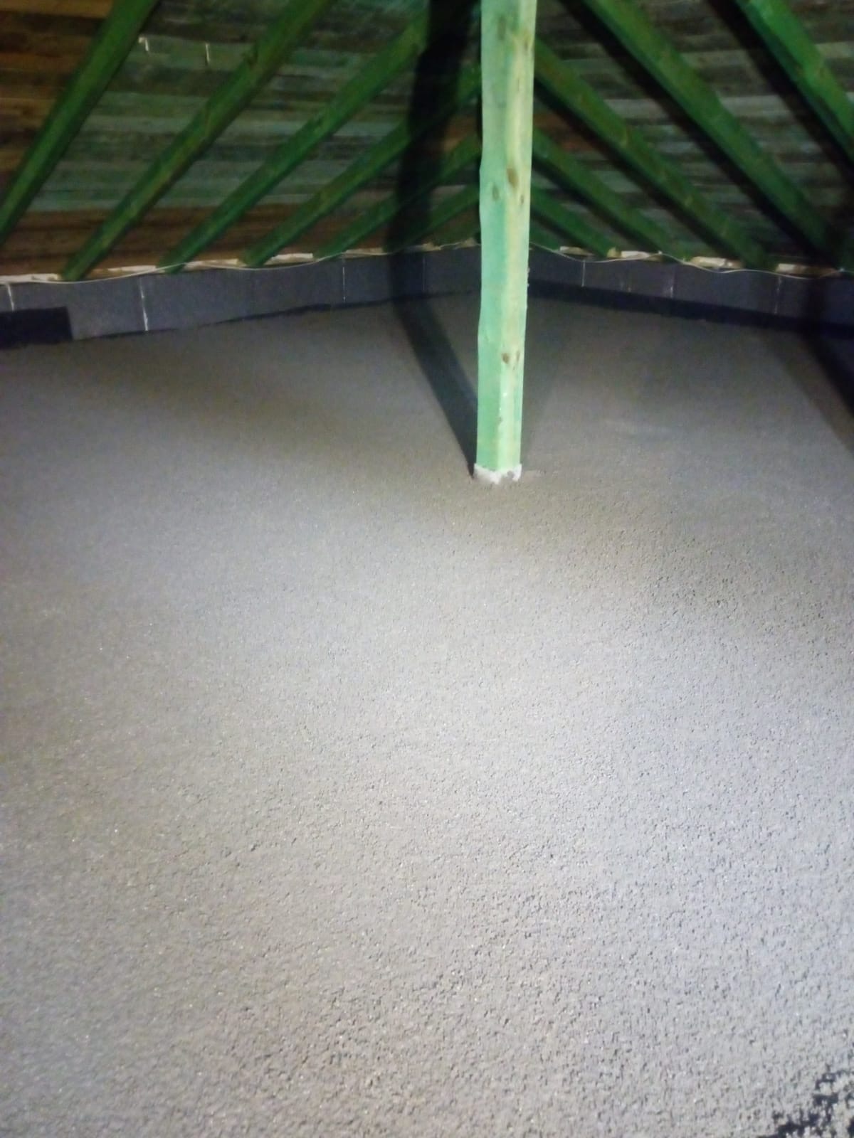 Świeżo wylana, gładka posadzka na poddaszu z widoczną drewnianą konstrukcją dachu i pionowym słupem wspierającym, otoczonym białym uszczelnieniem u podstawy.