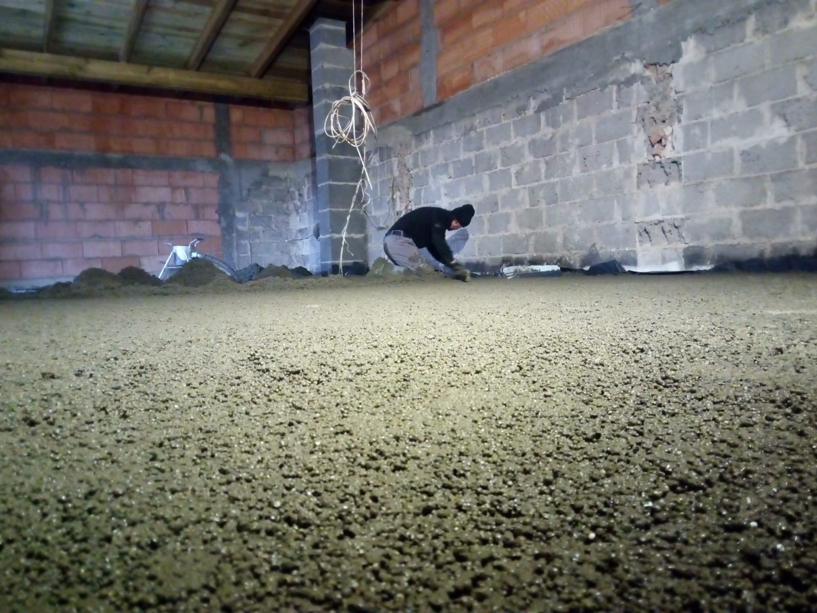 Pracownik wyrównuje świeżo wylaną, mokrą posadzkę w surowym wnętrzu z ceglanymi i betonowymi ścianami, widoczne przewody elektryczne zwisające z sufitu.