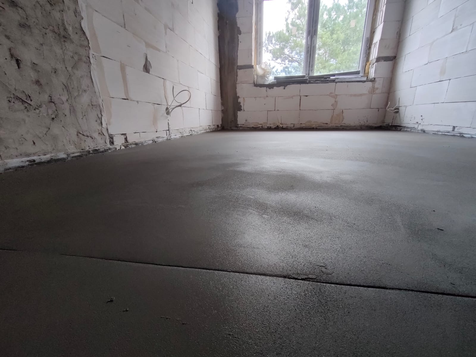 Świeżo wylana, gładka, szara posadzka w surowym wnętrzu z białymi ścianami z pustaków i oknem z widokiem na zieleń.