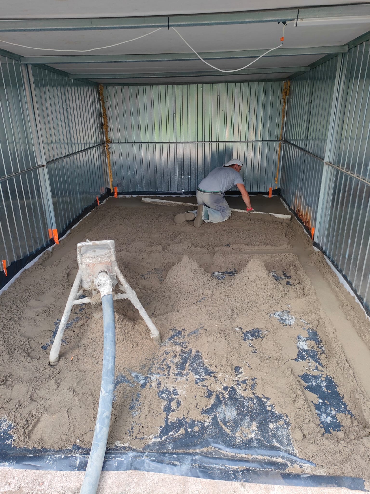 Pracownik wyrównuje mokrą zaprawę w blaszanym garażu, widoczna maszyna do podawania zaprawy i poziomice