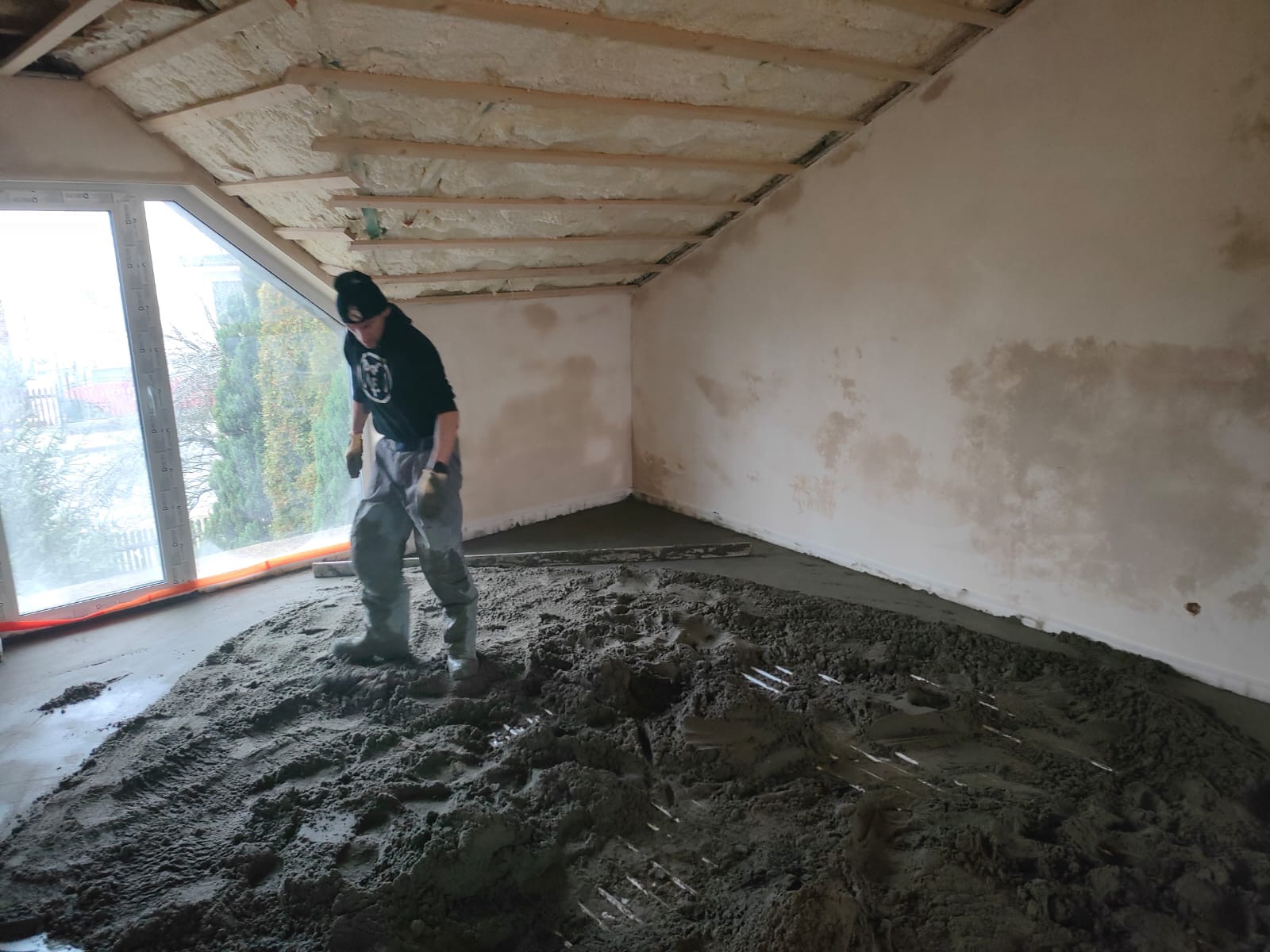 Pracownik w gumowych butach rozprowadza mokrą wylewkę na podłodze w pomieszczeniu z oknem i ocieplonym pianką sufitem, widoczne ślady butów i narzędzi w masie.