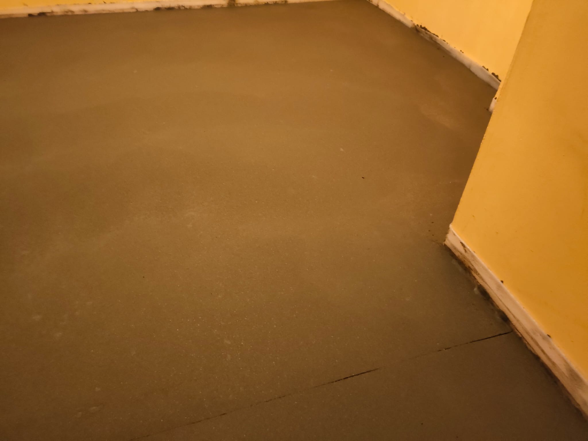 Świeżo wylana, gładka, szara posadzka w pomieszczeniu z żółtymi ścianami, widoczne łączenie w posadzce przy ścianie.