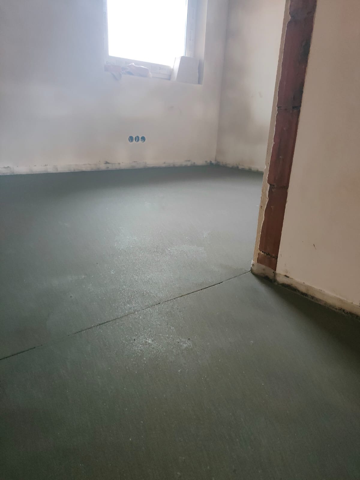 Świeżo wylana, wilgotna posadzka jastrychowa z widocznymi smugami i pęknięciami skurczowymi w surowym wnętrzu z oknem.