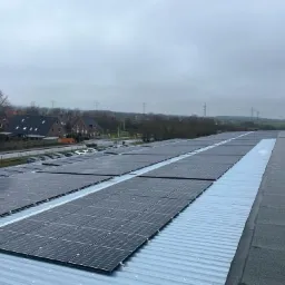 Rząd paneli słonecznych na płaskim dachu budynku przemysłowego, widoczne domy i linia wysokiego napięcia w oddali, pochmurne niebo.