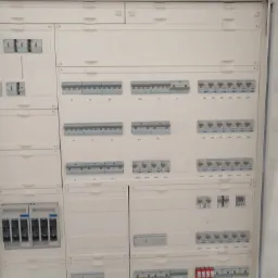 Rozdzielnica elektryczna z bezpiecznikami i wyłącznikami, widok z bliska, biała obudowa, oznaczenia na elementach.