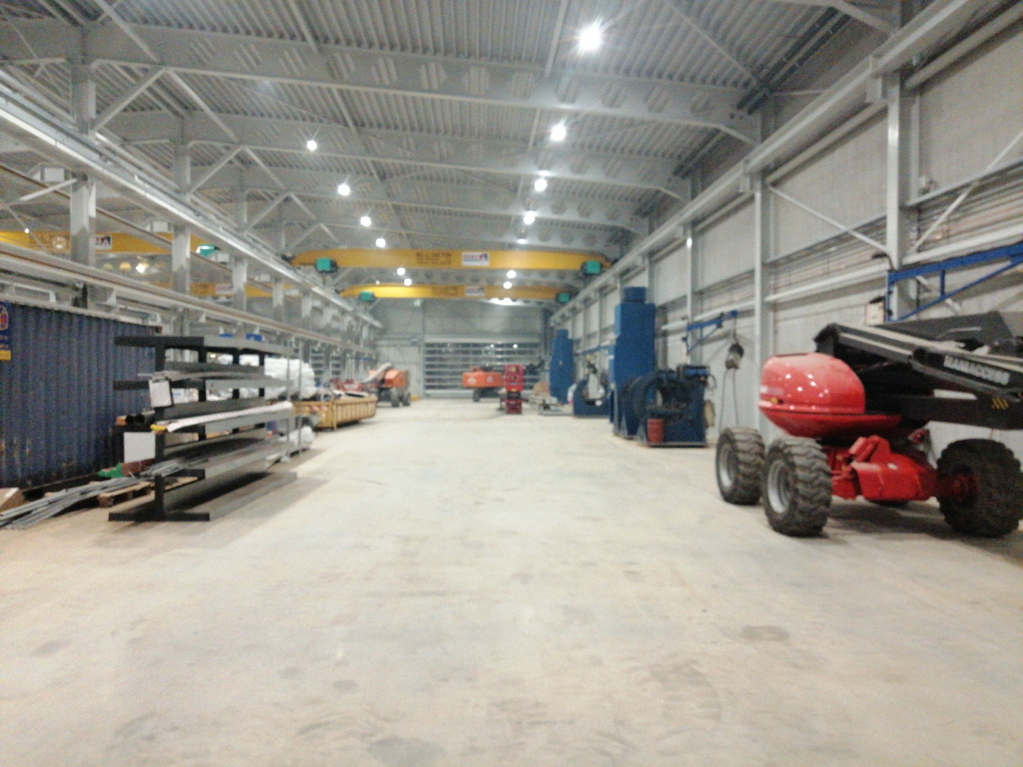 Wnętrze hali produkcyjnej z suwnicą, maszynami i materiałami budowlanymi, oświetlone lampami przemysłowymi.