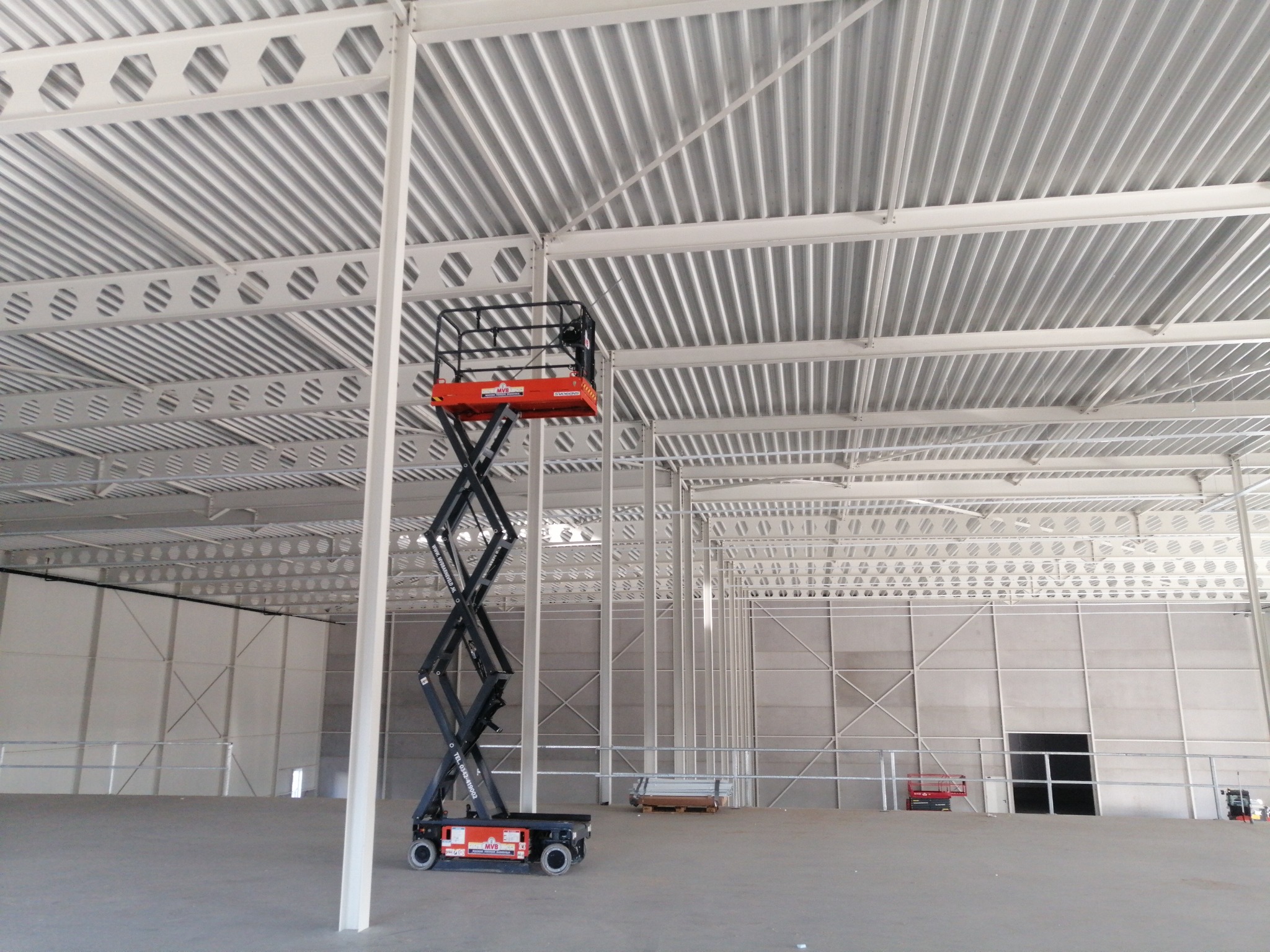 Wnętrze hali przemysłowej z podnośnikiem nożycowym na środku, widoczne białe stalowe konstrukcje dachu i ścian.