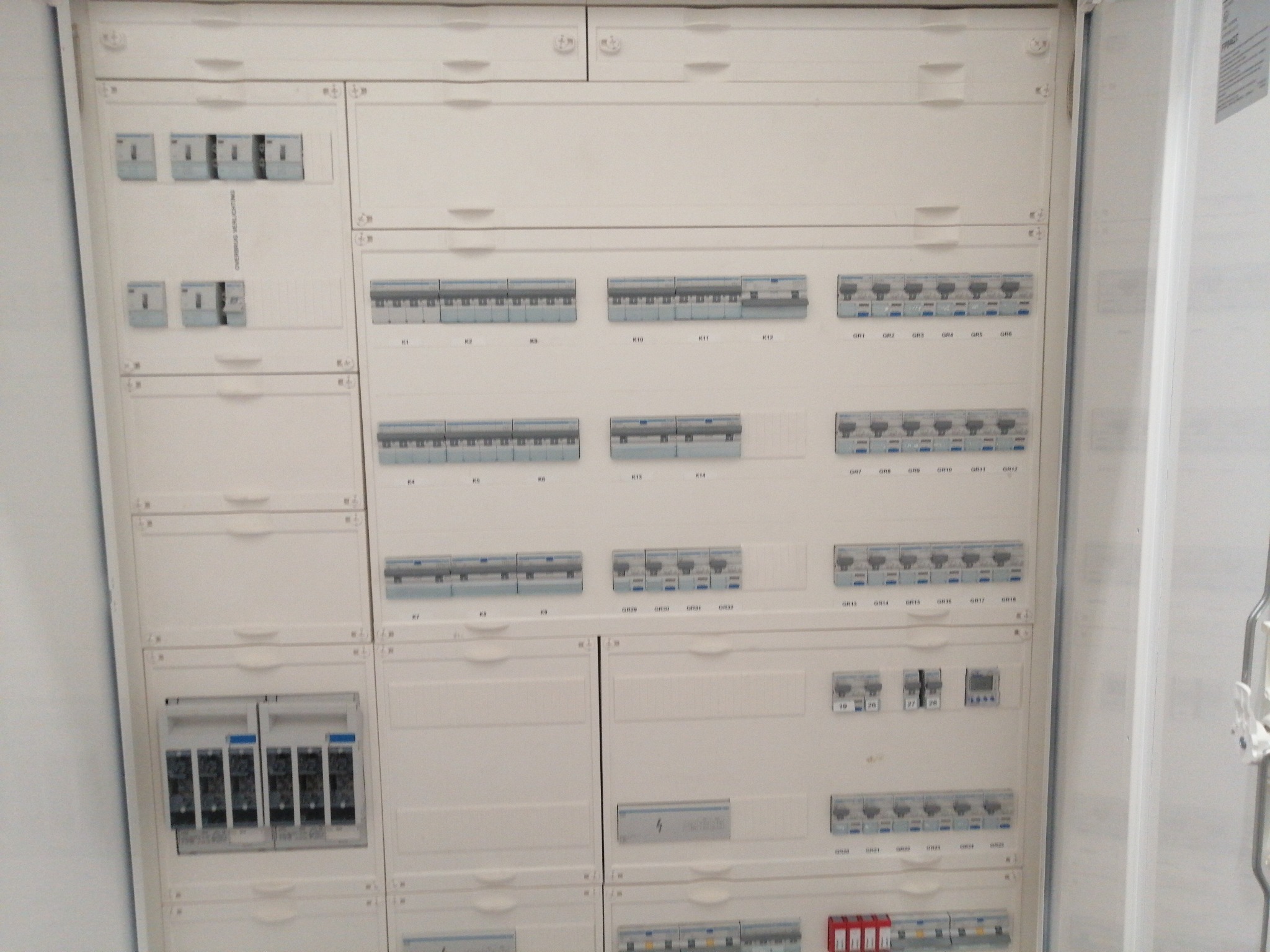 Rozdzielnica elektryczna z bezpiecznikami i wyłącznikami, widok z bliska, biała obudowa, oznaczenia na elementach.