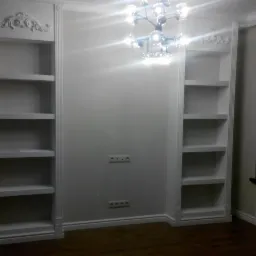 Białe, wbudowane regały z dekoracyjnymi elementami nad p&oacute;łkami, nowoczesny żyrandol z kulistymi kloszami i brązowa podłoga z paneli w pustym pokoju.