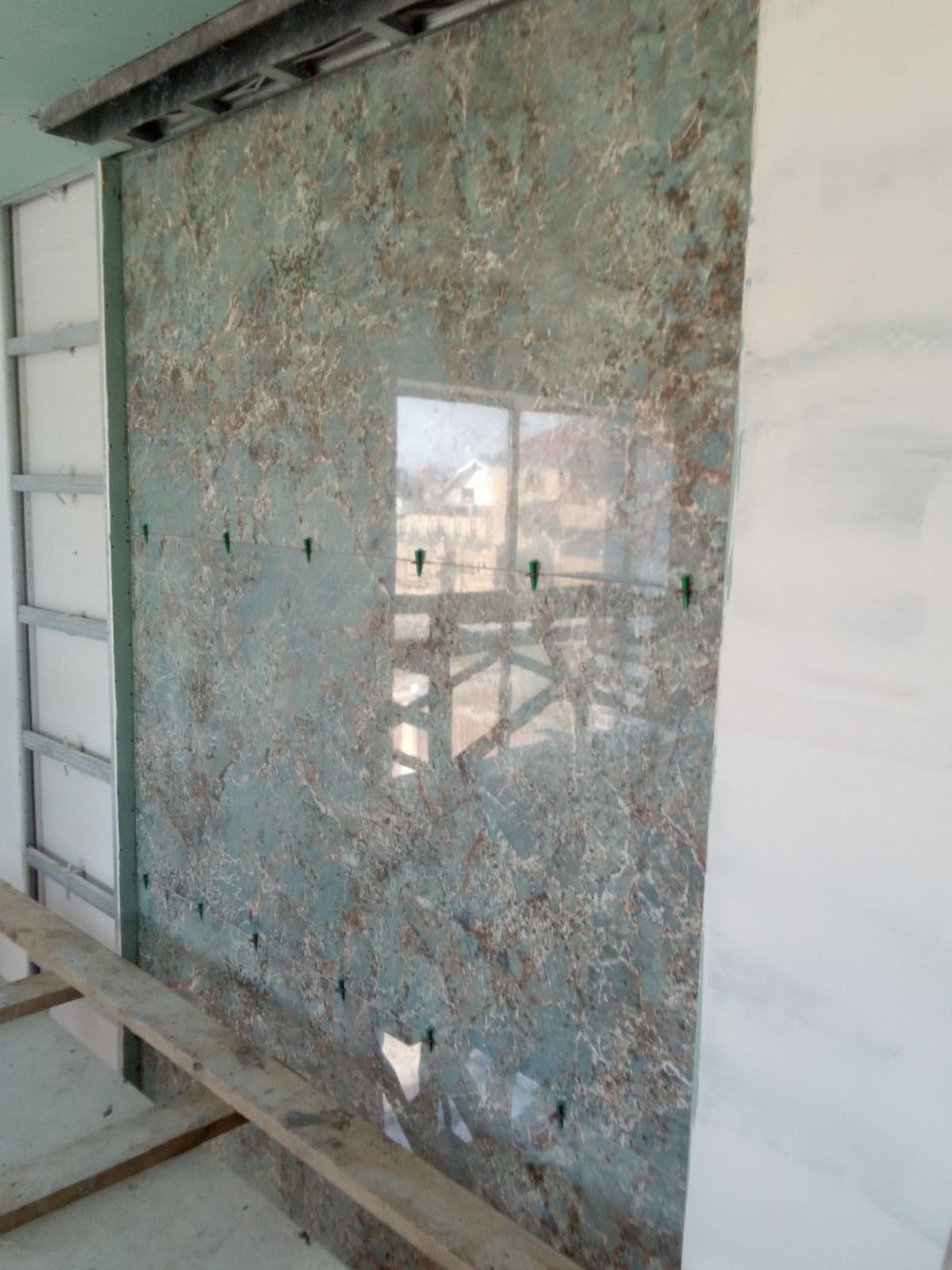 Ściana pokryta płytami z efektem marmuru w odcieniach szarości i brązu, zabezpieczona zielonymi klinami poziomującymi, widoczna konstrukcja metalowa i biała płyta gipsowa po bokach, odblask okna...