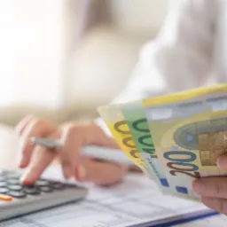 Osoba liczy pieniądze euro za pomocą kalkulatora, na biurku widoczne dokumenty i długopis.