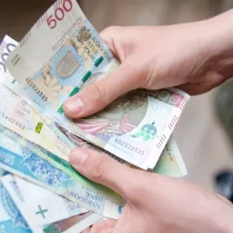Dłoń trzymająca plik banknotów polskich złotych o nominałach 500, 200 i 50 zł, z widocznymi detalami zabezpieczeń i numerami seryjnymi.