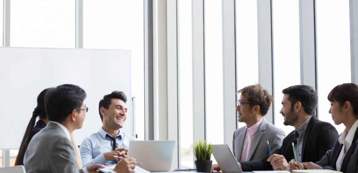 Zespół biznesowy omawia strategię przy stole konferencyjnym, skupiony na laptopach i dokumentach podczas spotkania w jasnym biurze z dużymi oknami.