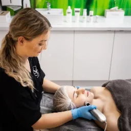 Kobieta w czepku poddawana zabiegowi kosmetycznemu na twarz przy użyciu urządzenia z metalową końcówką, wykonywanemu przez kosmetyczkę w niebieskich rękawiczkach. W tle widoczne kosmetyki i umywalka.