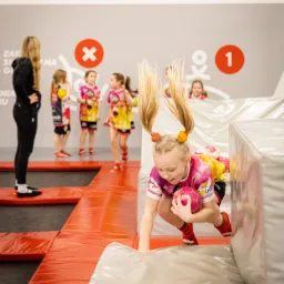 Dziewczynka z blond włosami spiętymi w dwa kucyki, w różowo-żółtej koszulce i czerwonych skarpetkach, z różową piłką w rękach, wspina się na miękką przeszkodę w sali zabaw Jum Now! w Gnieźnie.