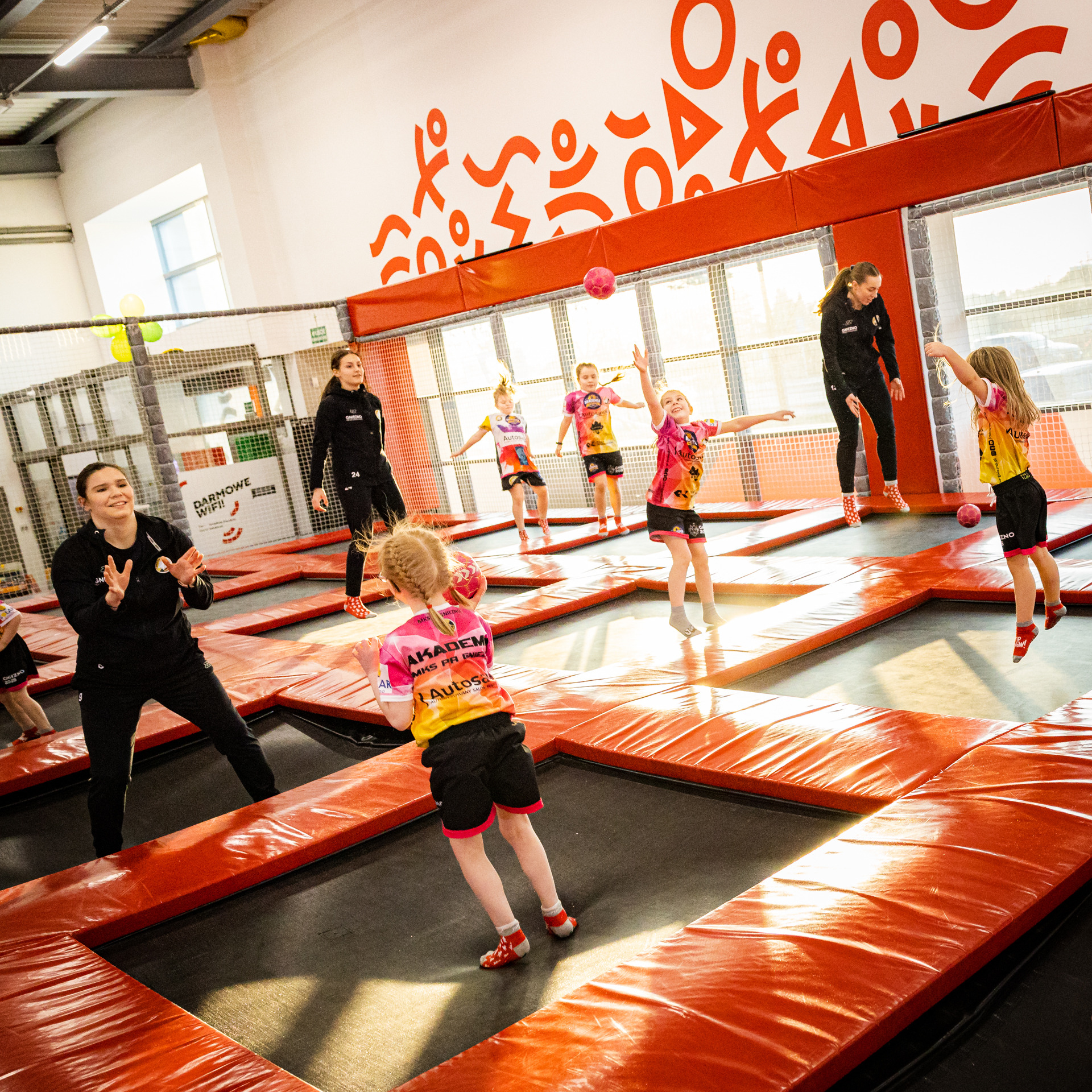 Grupa dziewcząt w sportowych strojach ćwiczy z piłkami na trampolinach pod okiem dwóch trenerek w jasnym, przestronnym wnętrzu sali gimnastycznej z pomarańczowymi akcentami.
